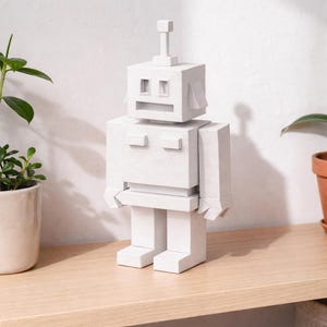 May include: A white, geometric robot sculpture with a rectangular head, body, and limbs. The robot has a small antenna and rectangular features for eyes and a mouth. It stands on a light-colored wooden surface.