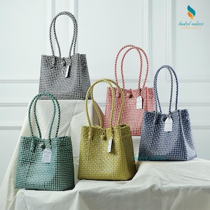 May include: A collection of woven tote bags in various colors, including green, yellow, red, blue, and black. Each bag features a braided handle and a tag. The bags are displayed on a white surface.