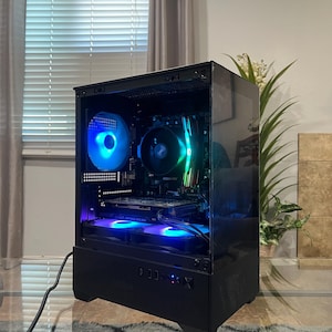 May include: A black desktop computer tower with a transparent side panel, featuring internal components illuminated by blue, green, and red LED lights. The case includes a front-facing power button, USB ports, and a mesh base design for ventilation.
