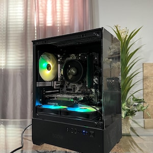 May include: A black desktop computer tower with a transparent side panel, featuring an internal motherboard, a central processor fan, and a Gigabyte graphics card. The case includes two illuminated multi-colored cooling fans at the base.