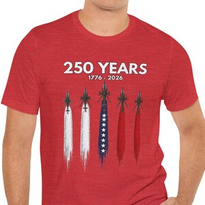 May include: A red short-sleeve t-shirt featuring the white text 250 YEARS 1776 - 2026 above a graphic of five fighter jets. The jets leave trails in white, blue with white stars, and red, mimicking the colors of the American flag.