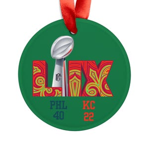 May include: A green round ornament with a red ribbon. The ornament features the Super Bowl LIX logo with the Vince Lombardi Trophy, red and green decorative lettering, and the scores PHL 40 and KC 22.