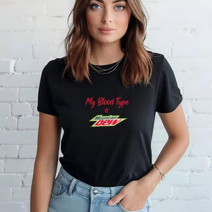 Puede incluir: Camiseta negra con el texto "My Blood Type is Mountain Dew" en rojo y blanco. El logotipo de Mountain Dew es amarillo, verde y rojo.