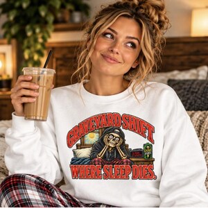 May include: A white crewneck sweatshirt featuring a graphic of a skeleton sitting in bed with the text "GRAVEYARD SHIFT" and "WHERE SLEEP DIES." The design includes a digital alarm clock showing 3:00 and a green energy drink can.