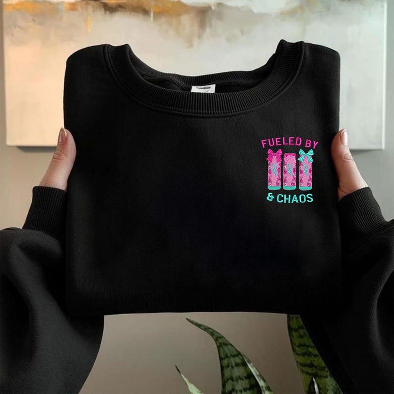 May include: Black sweatshirt with the text "Fueled by & Chaos" in pink and teal. The design features three pink and teal gift boxes with bows. The sweatshirt is held by a person.