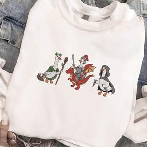 May include: White sweatshirt featuring embroidered cartoon geese in fantasy attire. One goose holds a staff, another is a knight riding a red dragon, and the third is hooded and armed with a knife. The design is whimsical and playful.