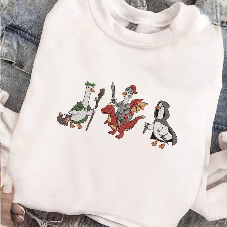 May include: White sweatshirt featuring embroidered cartoon geese in fantasy attire. One goose holds a staff, another is a knight riding a red dragon, and the third is hooded and armed with a knife. The design is whimsical and playful.