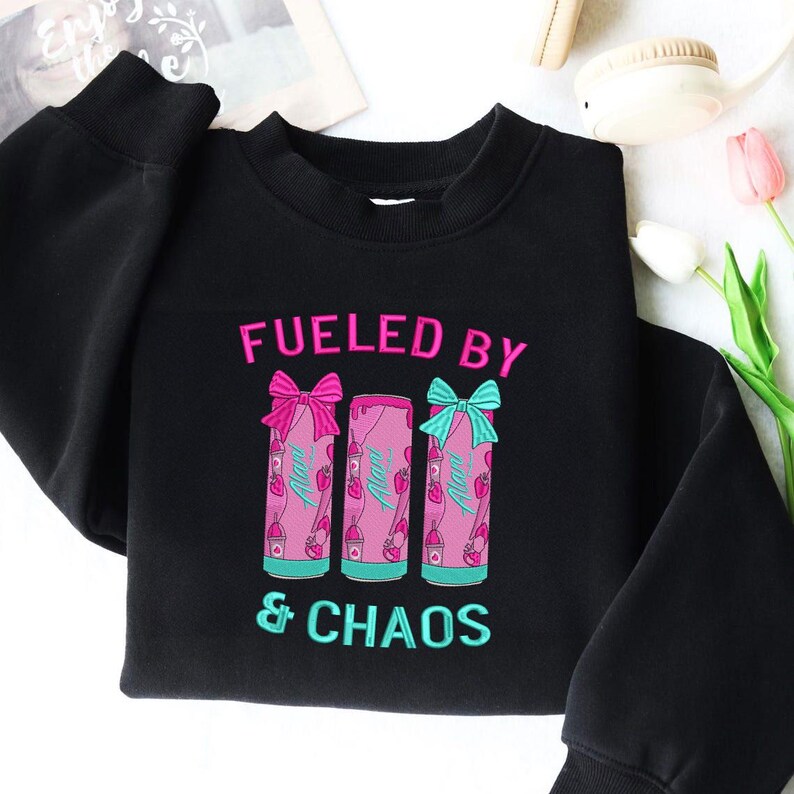 May include: Black sweatshirt with the embroidered text "FUELED BY & CHAOS." The design features three pink beverage cans with bows, strawberries, and the word "Alani." The text and graphics are in shades of pink and teal.