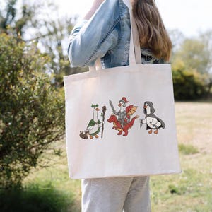 May include: Cream-colored tote bag featuring three cartoon characters: a goose with a staff, a knight riding a red dragon, and a penguin with a knife. The bag is carried by a person, with a blurred outdoor background.