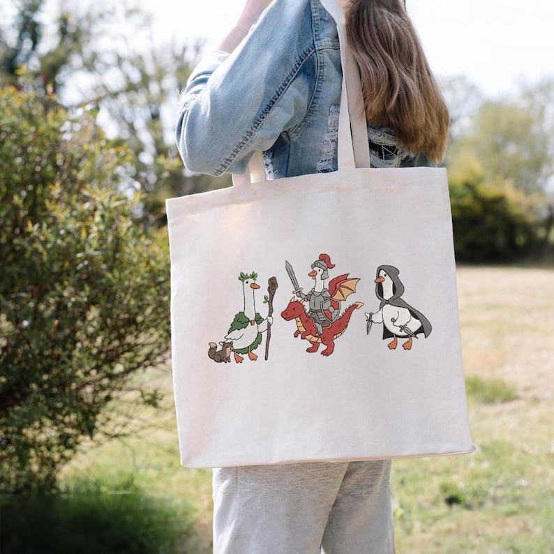 May include: Cream-colored tote bag featuring three cartoon characters: a goose with a staff, a knight riding a red dragon, and a penguin with a knife. The bag is carried by a person, with a blurred outdoor background.
