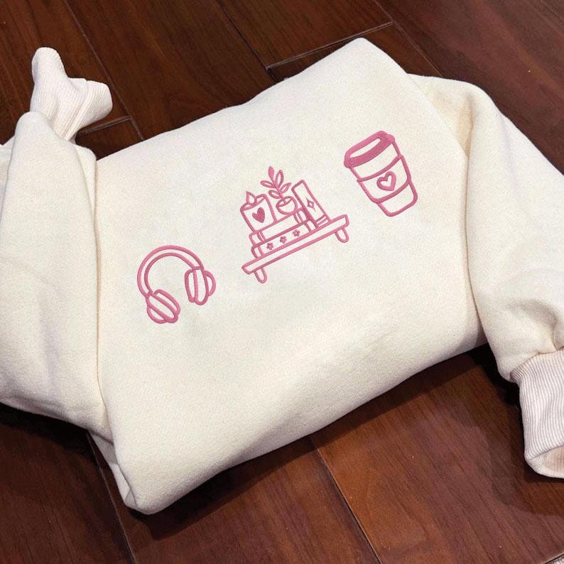 May include: Cream-colored sweatshirt with pink graphic icons of headphones, a bookshelf, and a coffee cup with a heart. The sweatshirt has a ribbed cuff and a relaxed fit, perfect for casual wear.