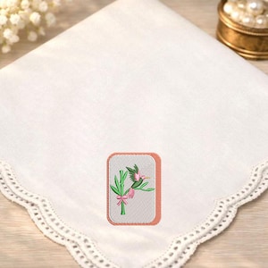 May include: A white square handkerchief featuring a scalloped lace edge and a central rectangular embroidered patch. The patch displays a green bamboo stalk with a pink bow and a small green and pink hummingbird perched on the stem.