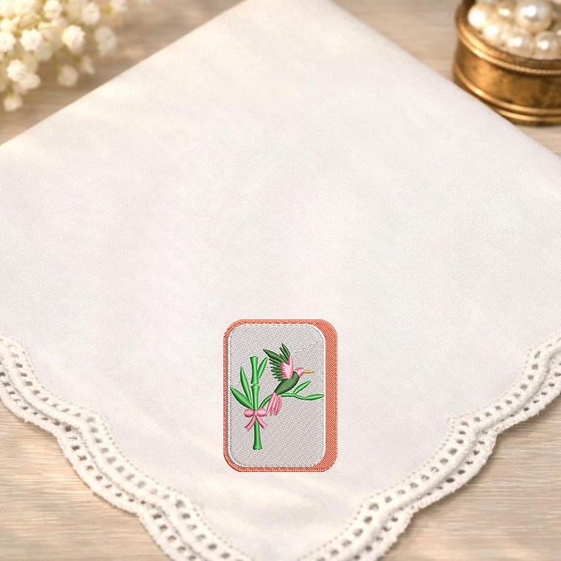 May include: A white square handkerchief featuring a scalloped lace edge and a central rectangular embroidered patch. The patch displays a green bamboo stalk with a pink bow and a small green and pink hummingbird perched on the stem.