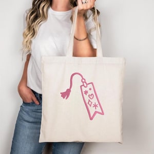 May include: A cream-colored tote bag with a pink embroidered design. The design features a tag with a tassel, hearts, diamonds, and stars. The bag has long handles and is held by a person.