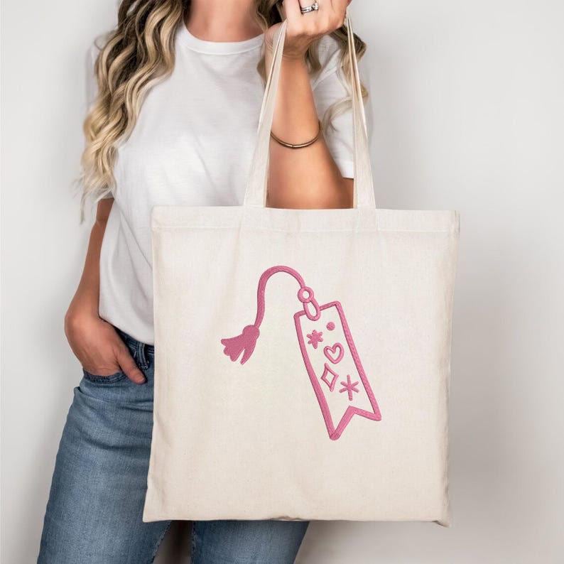 May include: A cream-colored tote bag with a pink embroidered design. The design features a tag with a tassel, hearts, diamonds, and stars. The bag has long handles and is held by a person.