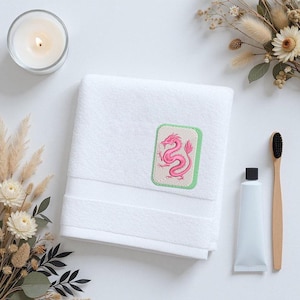 May include: A white cotton hand towel featuring a rectangular embroidered patch with a light green border. The patch contains a detailed pink dragon illustration on a beige background. The towel has a textured border near the bottom edge.