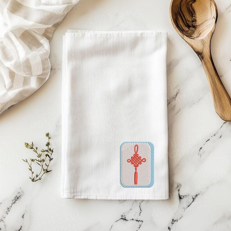 May include: A white rectangular kitchen towel featuring an embroidered patch in the bottom right corner. The patch has a light blue border, a beige background, and a red Chinese knot design with a tassel hanging from the bottom center.