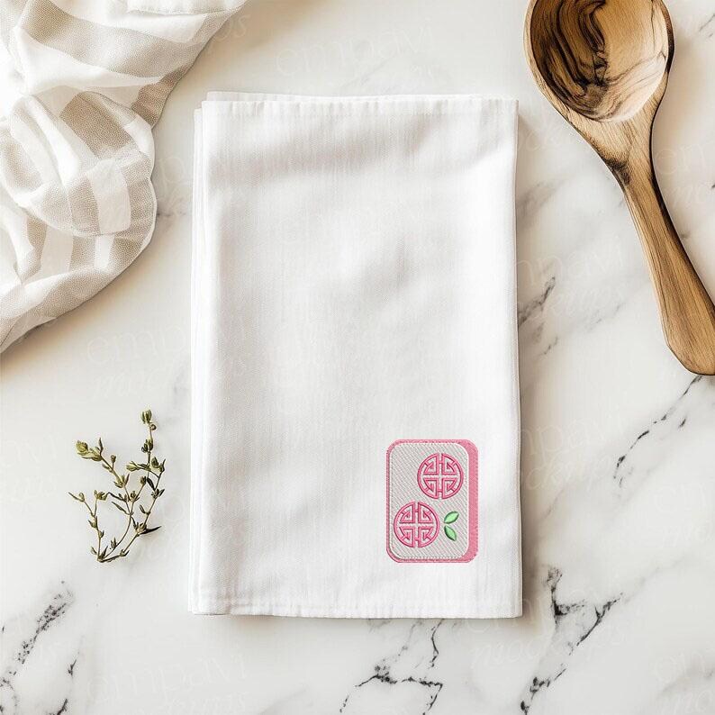 May include: A white rectangular kitchen towel featuring an embroidered graphic of a mahjong tile. The tile is light pink with a white center, displaying two circular pink symbols and a small green leaf accent in the bottom right corner.