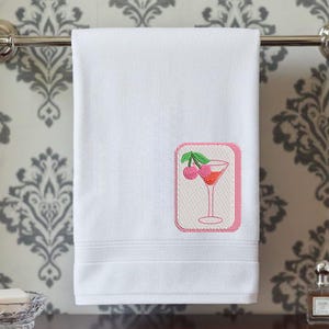 May include: A white hand towel featuring an embroidered rectangular patch with a pink border. The patch displays a martini glass filled with a reddish liquid and garnished with two pink cherries on a green stem, centered on the lower right side.