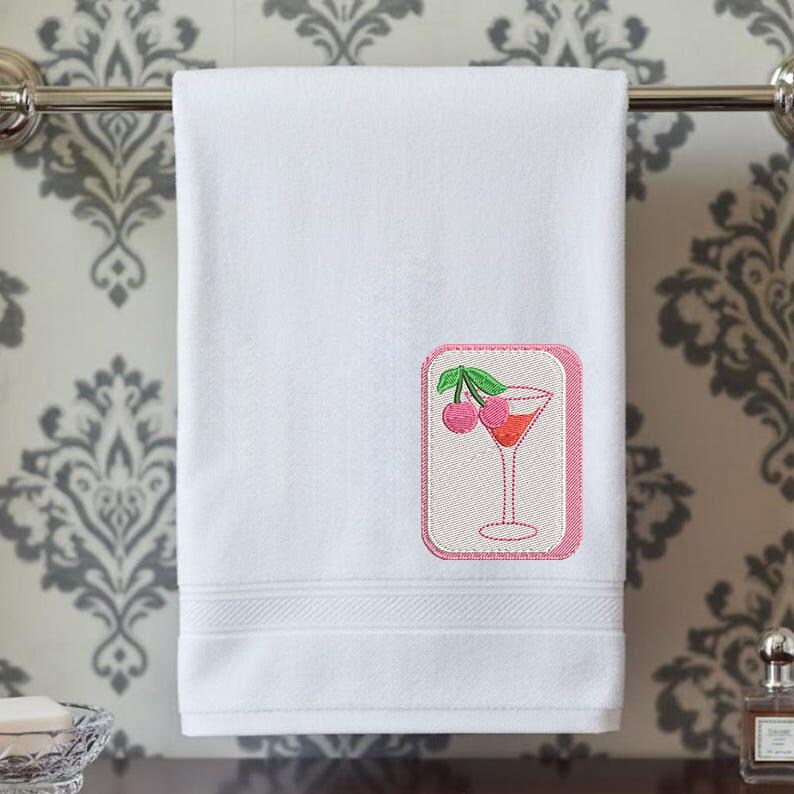 May include: A white hand towel featuring an embroidered rectangular patch with a pink border. The patch displays a martini glass filled with a reddish liquid and garnished with two pink cherries on a green stem, centered on the lower right side.