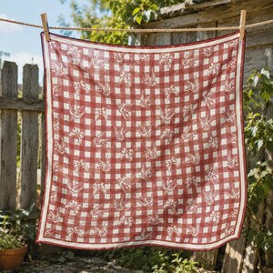 May include: A square scarf with a red and white gingham pattern, adorned with white bow and bird illustrations. The scarf is hanging on a clothesline with a wooden clothespin, showcasing a rustic aesthetic.
