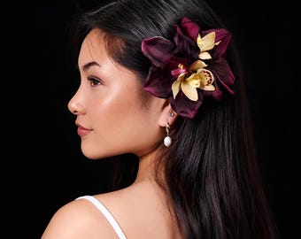 May include: A hair accessory featuring deep burgundy and cream-colored orchid flowers. The petals display a rich, dark purple hue with a contrasting pale yellow center, accented by subtle brown speckles on the inner lip of the floral bloom.