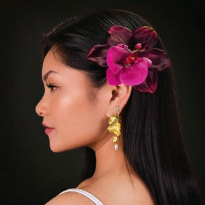 以下が含まれることがあります： 黒髪の女性が、髪に大きなマゼンタ色の蘭の花を飾っています。彼女はまた、真珠のドロップが付いた金色のイヤリングをしています。背景は無地の黒です。