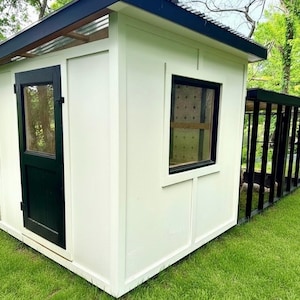 May include: A white outdoor structure with a black roof and trim, featuring a door and window with dark frames. The structure is set on a green lawn, with a black fence extending from the side.