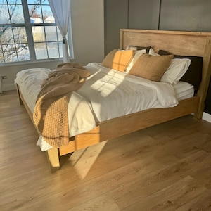 May include: A light wood bed frame with a tall headboard, featuring a white textured duvet, two mustard yellow square pillows, two white rectangular pillows, and a chunky knit mustard yellow throw blanket draped across the foot of the bed.