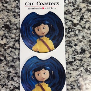 May include: Two round car coasters featuring a graphic of a character with blue hair wearing a yellow raincoat against a dark blue swirling background. The white display card above the coasters reads Car Coasters Handmade with love.