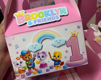 May include: A pink cardboard favor box featuring a white satin bow on top and the text "BROOKLYN & FRIENDS". The front displays a colorful illustration of cartoon animals, a rainbow, clouds, stars, and a large number one on a pedestal.
