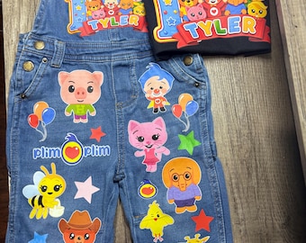 May include: A blue denim overall and a black t-shirt featuring Plim Plim circus-themed graphics. The overall displays cartoon animals, stars, and the text Plim Plim. The t-shirt shows a circus tent, the number 1, and the name TYLER in colorful letters.