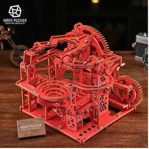Könnte beinhalten: Ein rotes 3D-Puzzle aus Holz mit einer Murmelbahn und Zahnrädern. Das Puzzle ist zusammengebaut und verfügt über eine Spiralbahn und eine Reihe von Zahnrädern, die die Murmeln auf der Bahn bewegen. Das Puzzle wird von Wood Puzzlea hergestellt.