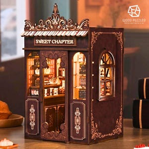 Puede incluir: Una tienda de madera en miniatura con el texto "SWEET CHAPTER" sobre la entrada. La tienda es marrón con intrincados diseños recortados y un interior iluminado, que muestra estantes en miniatura. El tema general es un modelo decorativo detallado.