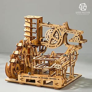 May include: A wooden marble run puzzle with gears, ramps, and a spiral lift. The natural wood-colored construction features multiple tracks for the marbles to travel through. The logo "WOOD PUZZLEA" is visible in the upper right corner.