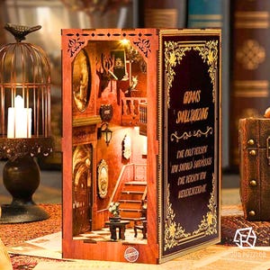 Puede incluir: Un rincón de libro de madera con una escena interior de una escalera, una mesa y una lámpara. El rincón del libro tiene un panel frontal decorativo con detalles dorados y texto. La escena está iluminada con pequeñas luces. Es un artículo decorativo.