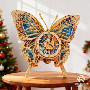 Puede incluir: Un reloj de mariposa de madera con intrincados detalles recortados. La esfera del reloj es redonda con números romanos. Las alas de la mariposa tienen detalles en azul y naranja. El reloj está sobre una mesa de madera.