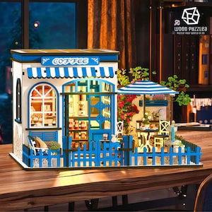 Puede incluir: Maqueta de cafetería de madera en miniatura, con una combinación de colores azul y blanco. El modelo presenta un letrero que dice "COFFEE", un toldo a rayas y una pequeña zona de estar al aire libre con una sombrilla a rayas.