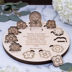 May include: Wooden prayer puzzle featuring a central figure and flower-shaped pieces. The puzzle includes the text of the Hail Mary prayer. The pieces are light brown with etched details, set on a round wooden base.