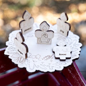 May include: A wooden centerpiece with five candle-shaped ornaments. Each candle has a dark brown flame and a word: hope, peace, joy, and love. The center candle has a baby in a basket. The centerpiece sits on a white, scalloped base.