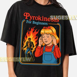 May include: Black t-shirt featuring a retro-style illustration of a girl with her eyes closed and hands on her temples. The illustration is surrounded by a blue and red border and includes the text "Pyrokinesis For Beginners".