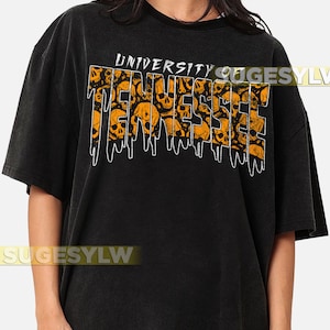 以下が含まれることがあります： オレンジ色の「UNIVERSITY TENNESSEE」の文字がスカルグラフィックで埋め尽くされた黒いTシャツ。テキストは白いアウトラインで滴るような効果があります。シャツは半袖でクルーネックです。