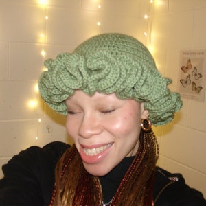May include: A green crocheted hat with a ruffled design. The hat is worn by a person with long brown and red braids.