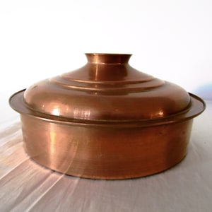 May include: A copper-coloured, round lidded pot. The pot has a slightly flared base and a domed lid with a small, cylindrical handle. The metal has a warm, reddish-brown hue and a polished finish. The pot is likely used for cooking or serving.