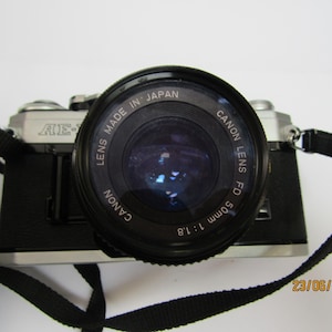 Puede incluir: Cámara Canon AE-1 negra y plateada. La lente es negra y tiene la inscripción "Canon Lens FD 50mm 1:1.8 Made in Japan". Una correa negra está adjunta. La fecha 23/06/2025 es visible.