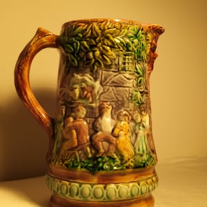 May include: A brown ceramic pitcher with a green and brown glaze. The pitcher features a raised design of people and buildings. The pitcher has a handle and a wide mouth.