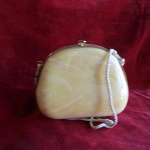 Puede incluir: Un bolso de mano blanco y amarillo con efecto marmoleado, cierre dorado y cadena blanca.