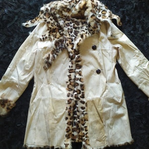May include: A cream-coloured leather jacket with a leopard print fur collar and trim. The jacket has a double-breasted closure with two buttons.