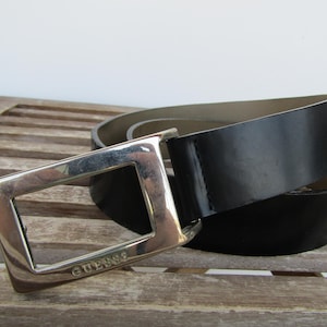 May include: A black belt with a shiny finish and a silver-colored square buckle. The buckle has the word "GUESS" engraved on it. The belt is lying on a wooden surface.