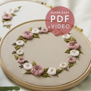 Puede incluir: Primer plano de un aro de bordado beige con un diseño de corona floral. La corona está hecha de rosas rosas y blancas y hojas verdes. El texto "SUPER EASY PDF + VIDEO" está impreso en blanco sobre un círculo rosa.